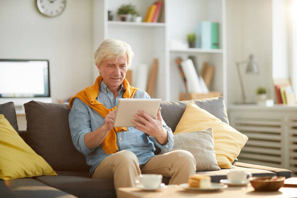 Portrait of modern senior man using digital tablet while relaxing sitting on comfortable couch at home, copy space