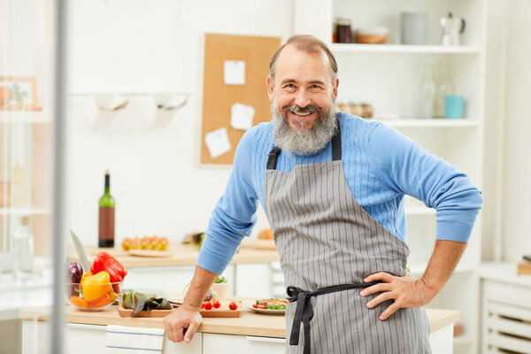 Waist up portrait of bearded senior man wearing apron looking at camera cheerfully while cooking at home, copy space