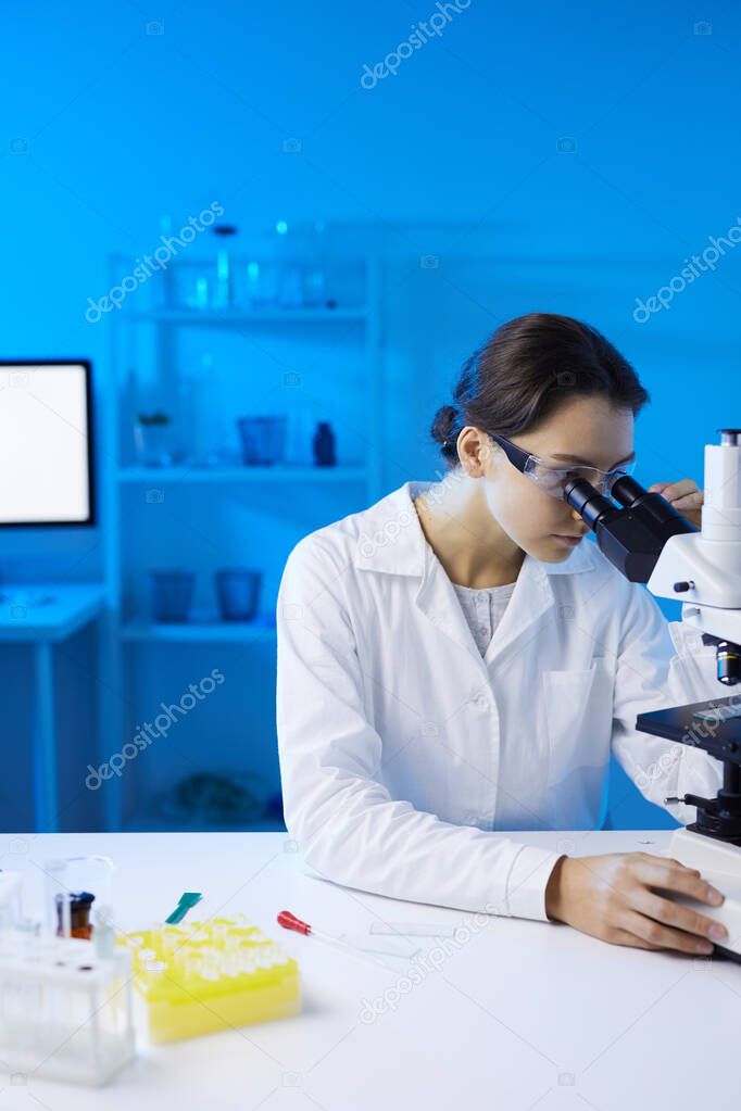 Portrait of young female scientist looking in microscope while doing research in medical laboratory lit by blue light, copy space