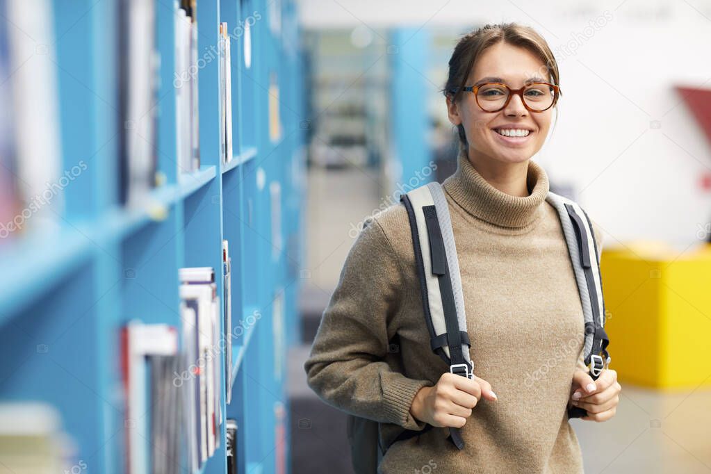 Waist up portrait of female college student wearing backpack and smiling at camera cheerfully while posing in library, copy space