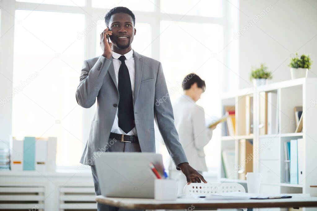 Portrait of smiling African-American businessman speaking by phone while standing by workplace in office, copy space
