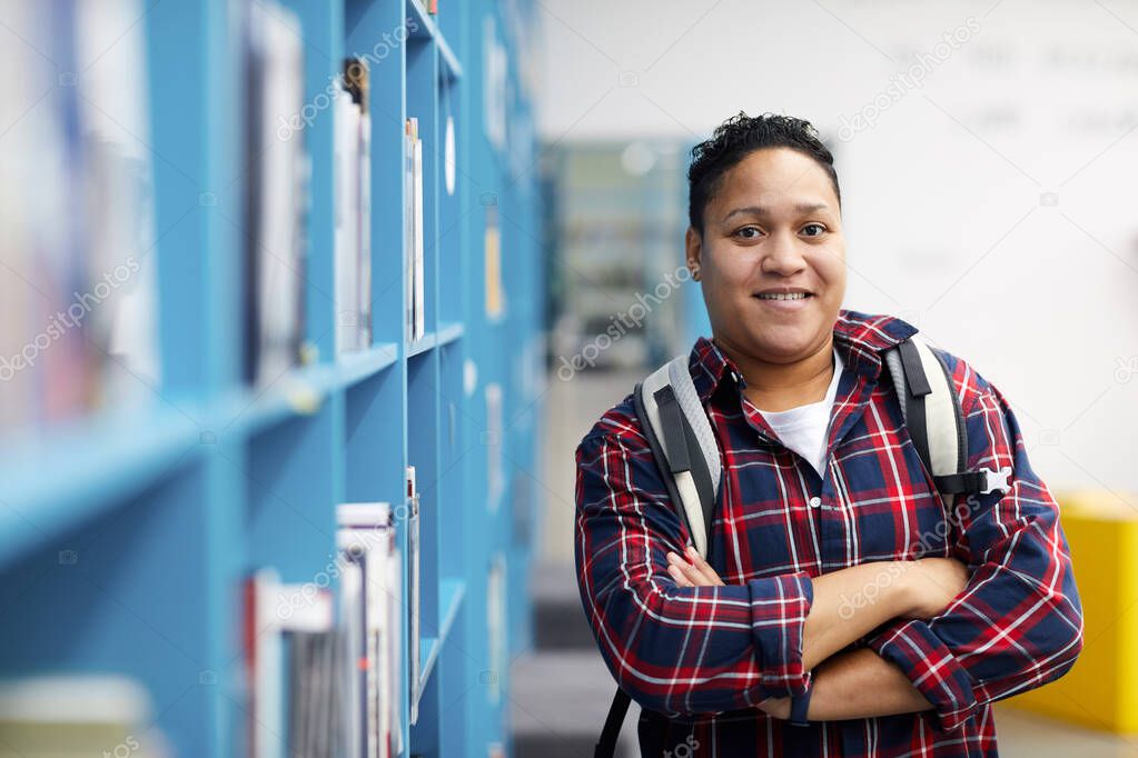 Waist up portrait of mixed-race college student wearing backpack and smiling at camera cheerfully while posing in library, copy space