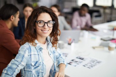 Yaratıcı ekiple toplantı masasında otururken kameraya bakan gülümseyen kadın tasarımcının portresi.