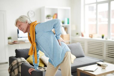 Sırt ağrısı çeken ve evdeki kanepeye yaslanan yaşlı bir adamın yan görünüm portresi.