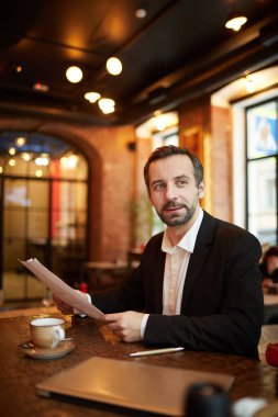 Yakışıklı, olgun bir iş adamının portresi lüks bir restoranda masada otururken ve elinde belge tutarken dalgın dalgın bakıyor.