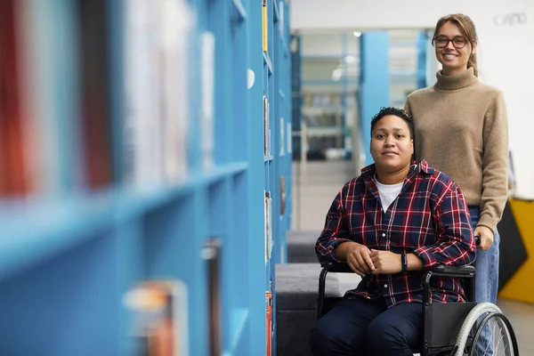 两个大学生的画像 其中一个坐在轮椅上 站在图书馆的架子旁边 复印空间 — 图库照片