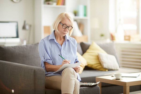Portrait of modern senior woman calculating finances and filling in forms while preparing tax report at home, copy space