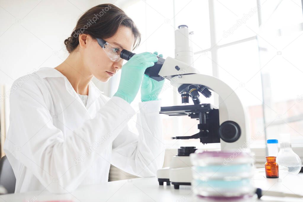 Side view portrait of young woman looking in microscope while working on research in medical laboratory, copy space