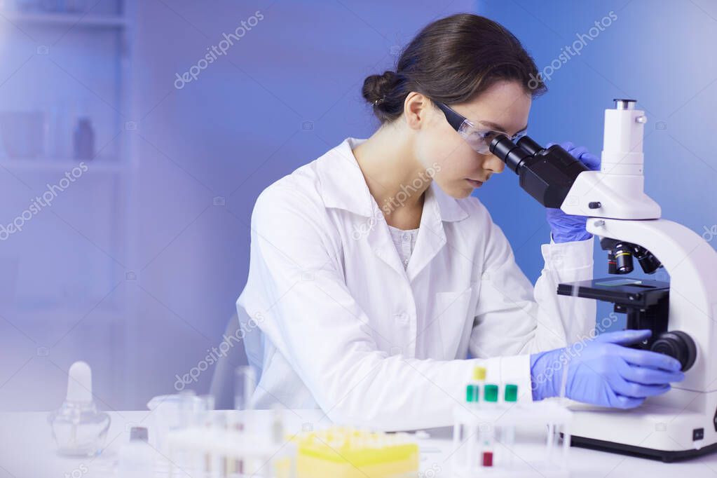 Side view portrait of beautiful young woman looking in microscope while working on medical research in laboratory, copy space