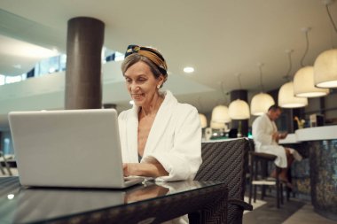 Portrait of mature woman smiling happily while using laptop in restaurant at luxury SPA resort, copy space