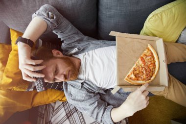 Above view portrait of bearded man lying on couch and eating pizza while watching TV at home, copy space clipart