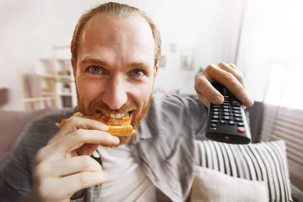 Fish eye portrait of smiling bearded man watching TV at home and eating pizza while binge watching favorite series, copy space