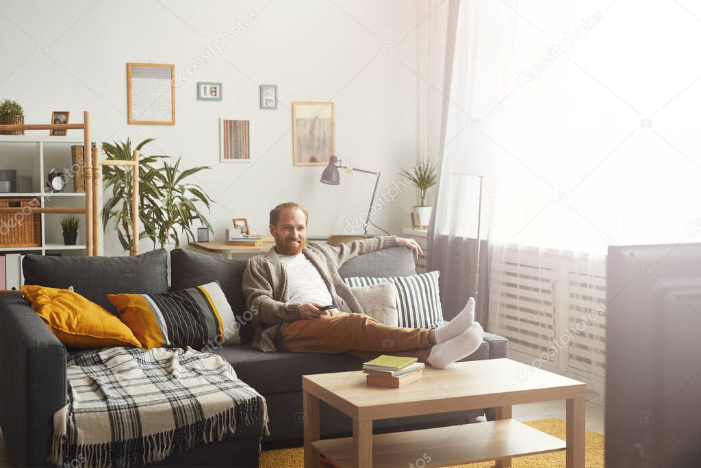 Full length portrait of modern adult man relaxing on couch while watching TV at home, copy space