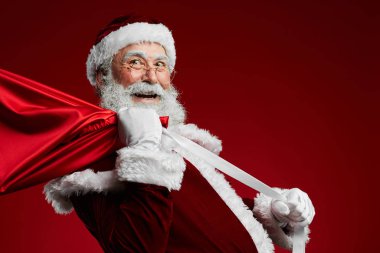 Waist up portrait of smiling Santa Claus holding bag with Christmas presents over red background, copy space clipart