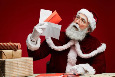 Elinde mektuplar olan klasik Noel Baba portresi kırmızı arka planda Noel hediyeleriyle masada oturuyor.