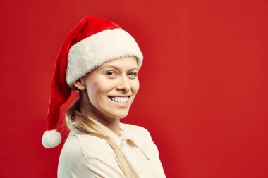 Sarışın, Noel Baba şapkası takan, kırmızı arka planda poz verirken mutlu bir şekilde gülümseyen genç kadın portresi.