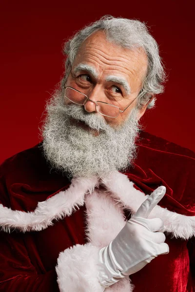Waist up portrait of classic Santa Claus shaking finger at someone and looking away while posing against red background