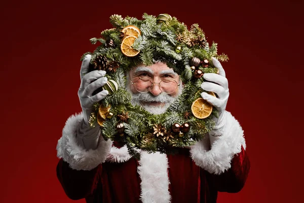 举起经典圣诞老人的画像 拿着圣诞花环 看着站在红色背景下的相机 — 图库照片