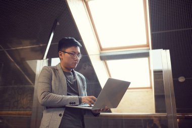 Modern ofis içinde dururken dizüstü bilgisayar kullanan başarılı Asyalı iş adamının düşük açılı portresi, boşluğu kopyala