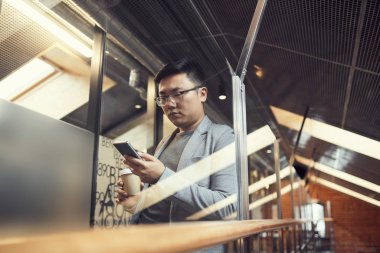 Modern ofis içinde cam duvarın arkasında duran akıllı telefon kullanan genç Asyalı işadamının belden yukarı portresi.