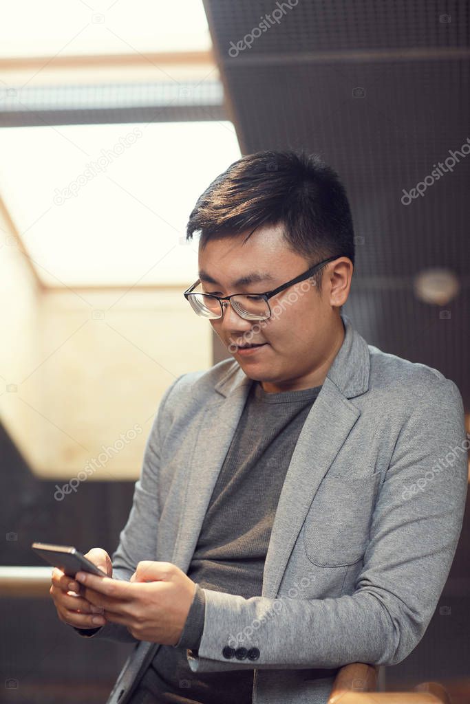 Waist up portrait of successful Asian man using smartphone while leaning on rail in modern office