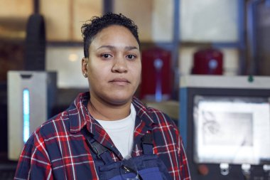 Melez kadın işçinin kafa ve omuz portresi atölyede endüstriyel makine ünitelerinin yanında duran kameraya bakıyor.