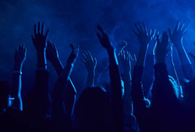 Mavi ışıkla aydınlatılan dumanlı gece kulübündeki müzik konserinin keyfini çıkarırken el kaldıran büyük bir grup insan silueti.