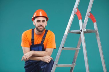 Tek bacağı merdivende dinlenen yakışıklı işçinin yatay olarak çekilmiş portresi.