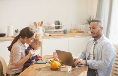 Modern babanın portresi dizüstü bilgisayar kullanırken sıcak mutfağın içinde aile kahvaltısının tadını çıkarıyor.
