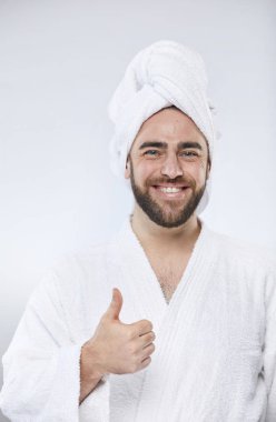 Havlu başlıklı ve bornozlu neşeli bir gencin stüdyo portresi dikey olarak başparmağıyla kameraya bakıp gülümsüyor.