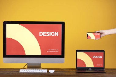 Web design concept with hand holding smartphone, computer and laptop at wooden table over pop yellow background, copy space