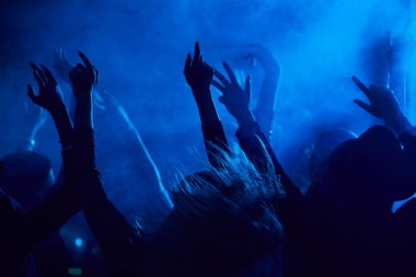 Mavi ışıkla aydınlatılan dumanlı gece kulübünde müzik konserinin keyfini çıkarırken zıplayan ve el kaldıran gençlerin siluetleri.