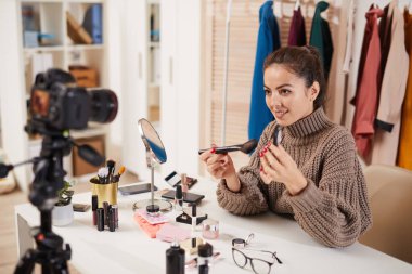 Makyaj ürünlerini test eden ve video incelerken kamerayla konuşan gülümseyen genç bir kadının portresi veya güzellik ve yaşam tarzı kanalı için özel ders, kopyalama alanı