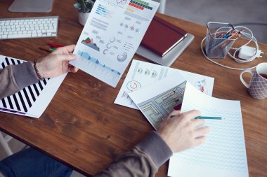 Closeup of unrecognizable modern businessman holding statistics report with colored data while working at wooden desk in office, copy space
