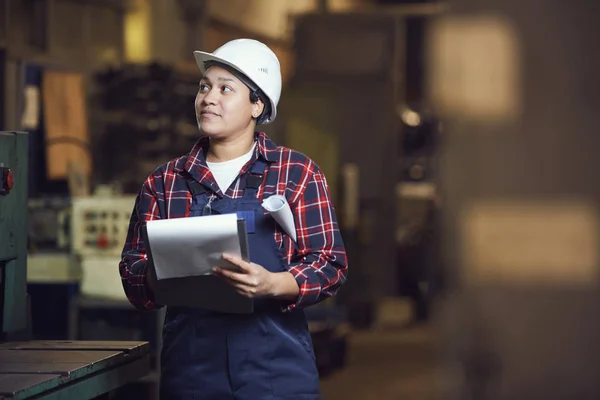 Waist up portrait of modern female worker holding clipboard while standing by machine units in industrial plant, copy space