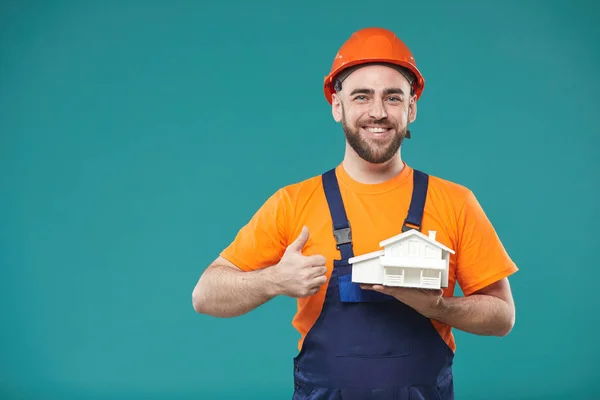 Horizontal studio portrait of construction worker posing on camera with dummy building standing against turquoise background