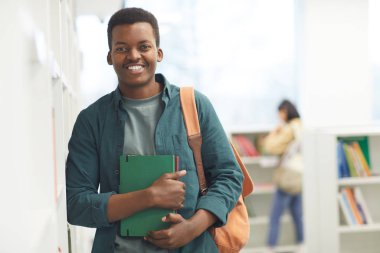Kolej kütüphanesinde raflarda poz verirken kameraya bakıp gülümseyen Afro-Amerikan öğrencinin belden yukarı portresi.