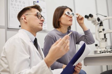 Modern oftalmoloji kliniğinde görüş testi sırasında genç bir kadınla konuşan genç optometristin yan görüş portresi, fotokopi alanı