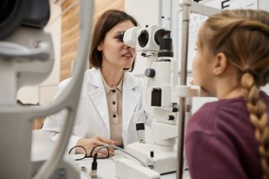 Modern klinikte küçük bir kızın görme mesafesini kontrol ederken kırılma ölçerini kullanan kadın göz doktorunun portresi.