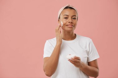 Waist up portrait of Mixed-Race young woman applying face cream and smiling at camera while posing against pink background during beauty routine in morning clipart