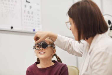 Modern oftalmoloji kliniğinde küçük bir kızın görüşünü kontrol ederken, deneme çerçevesi oluşturan kadın göz doktorunun portresi.