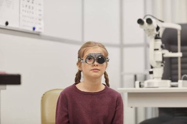 Portrait of cute little girl wearing trial frame during vision test in pediatric ophthalmology clinic, copy space clipart