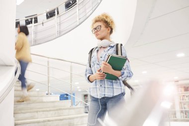 Modern üniversitenin iç kısımlarında, sarmal merdivenlerden inerken elinde kitap tutan kız öğrencinin belden yukarı portresi.