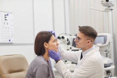 Tıbbi klinikte vizyonunu kontrol ederken kadın hastanın gözünü açan genç göz doktorunun yan görüş portresi.
