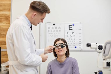 Portrait of male optometrist putting trial frame on female patient during vision check in modern clinic, copy space clipart