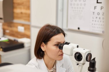 Modern klinikte göz testi sırasında refraktometre kullanan genç kadın göz doktorunun yüksek açılı portresi.
