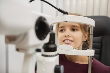 Portrait of cute smiling girl looking into refractometer during vision test in modern ophthalmology clinic, copy space clipart