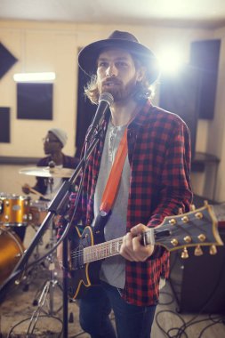 Çağdaş, uzun saçlı, gitar çalan ve müzik grubuyla prova ya da konser sırasında mikrofona şarkı söyleyen adamın belden yukarı portresi