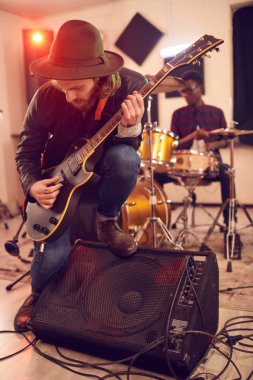 Çağdaş genç adamın tam boy portresi. Elektro gitar solosu çalıyor. Konser sırasında ya da provalar sırasında arka planda müzik bandıyla amfiye yaslanıyor.