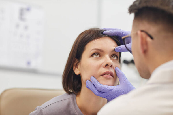 Back view portrait of young ophthalmologist opening eye of female patient while checking her vision in med clinic, copy space